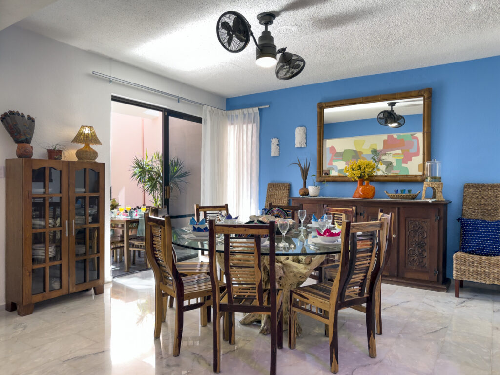 Glass-topped dining table, wooden chairs, and sideboard beneath a large mirror on a blue wall.