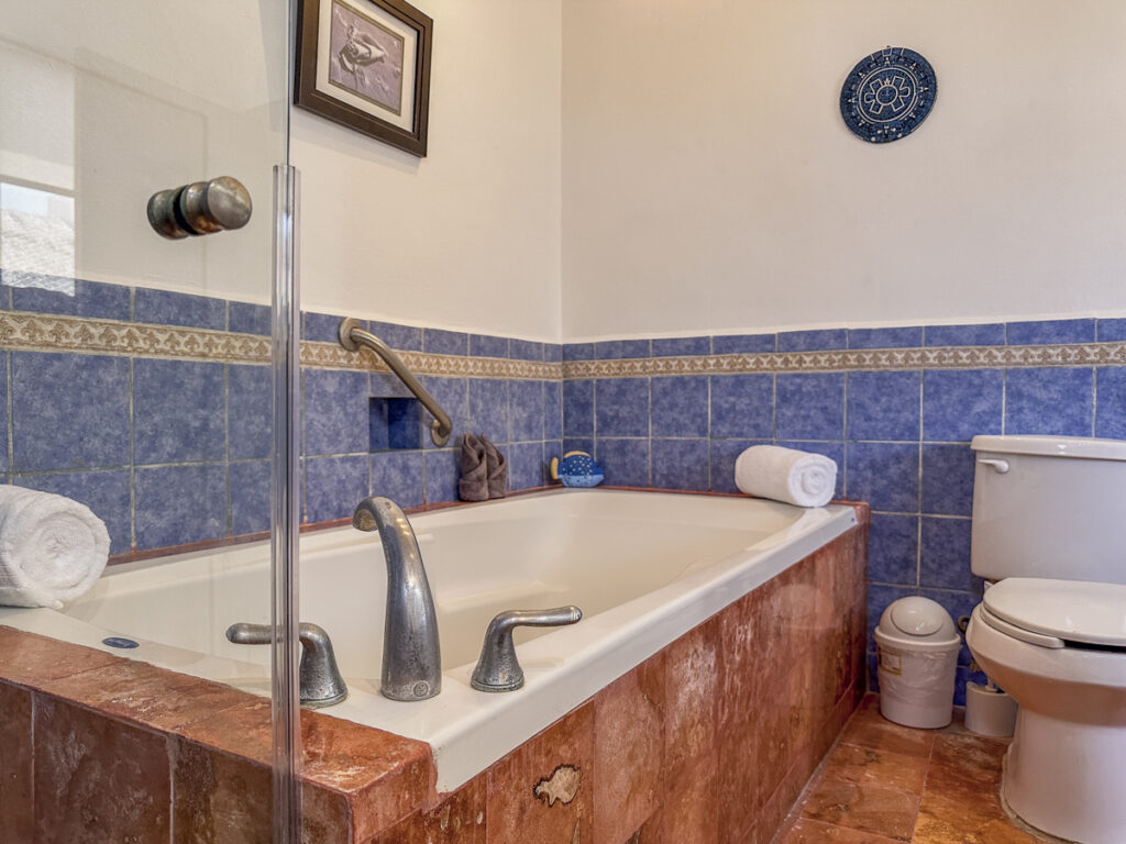 Bathtub with handrails beside a toilet and blue-bordered tiled walls.