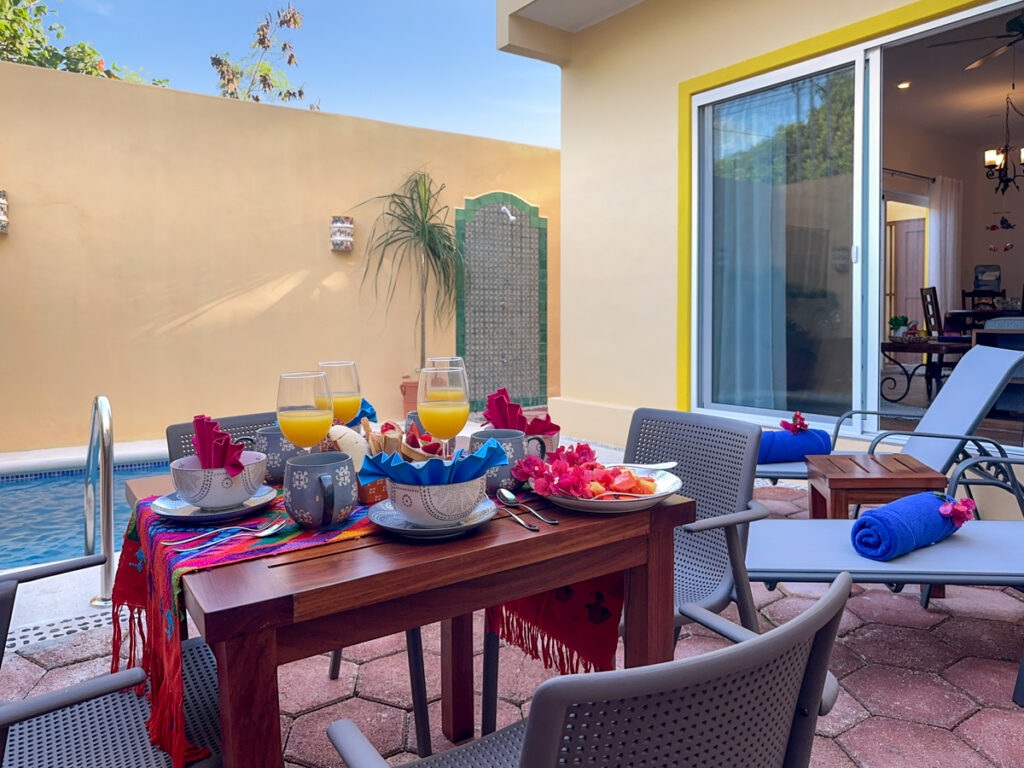 Patio table set with dishes, drinks, and colorful napkins beside lounge chairs and a sliding door.