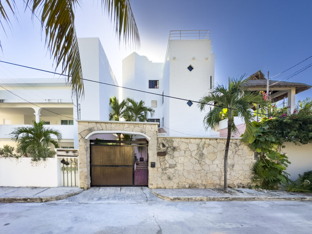 Tall white house behind a stone wall and arched wooden gate beneath palms and blue sky.
