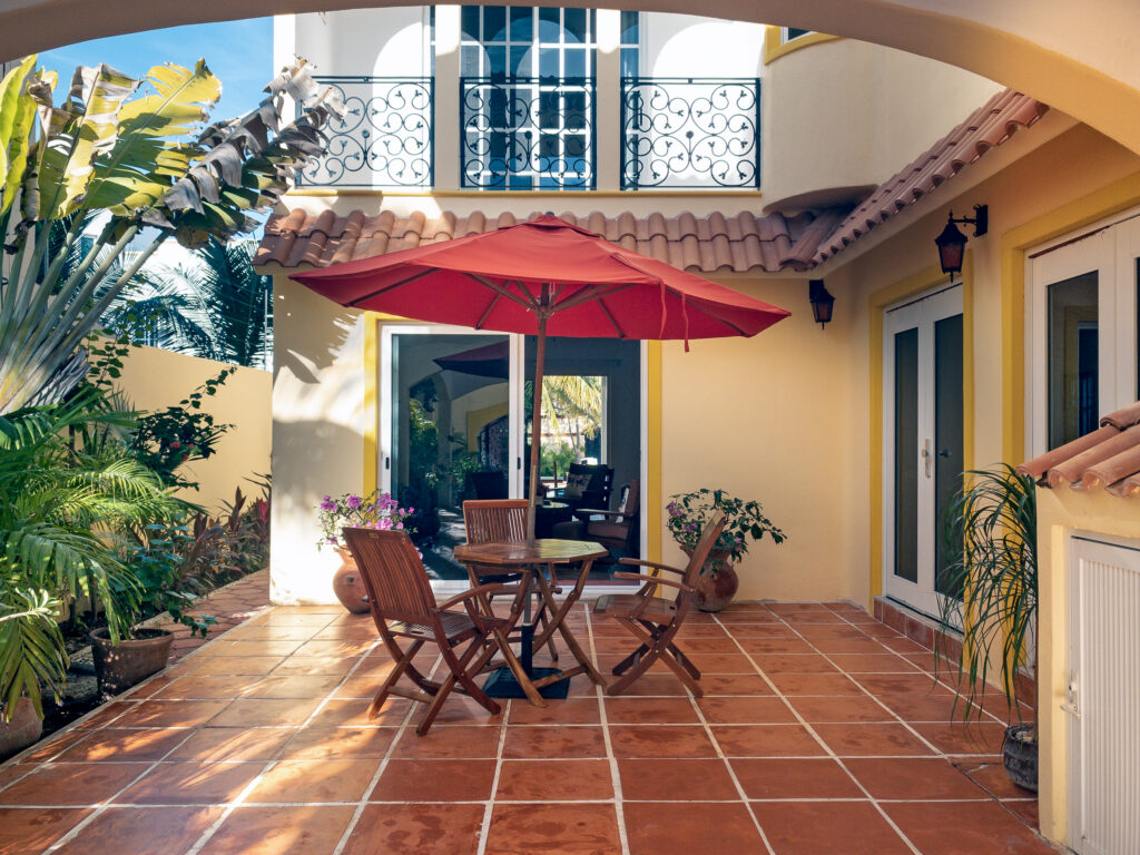 Wooden table and chairs beneath a red umbrella in a tiled courtyard surrounded by the house.