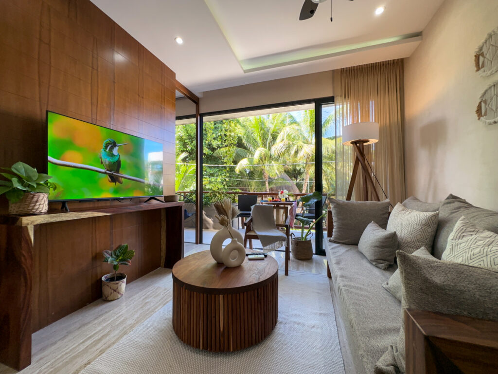 Gray sofa and round wooden coffee table facing a television beside glass balcony doors overlooking palms.