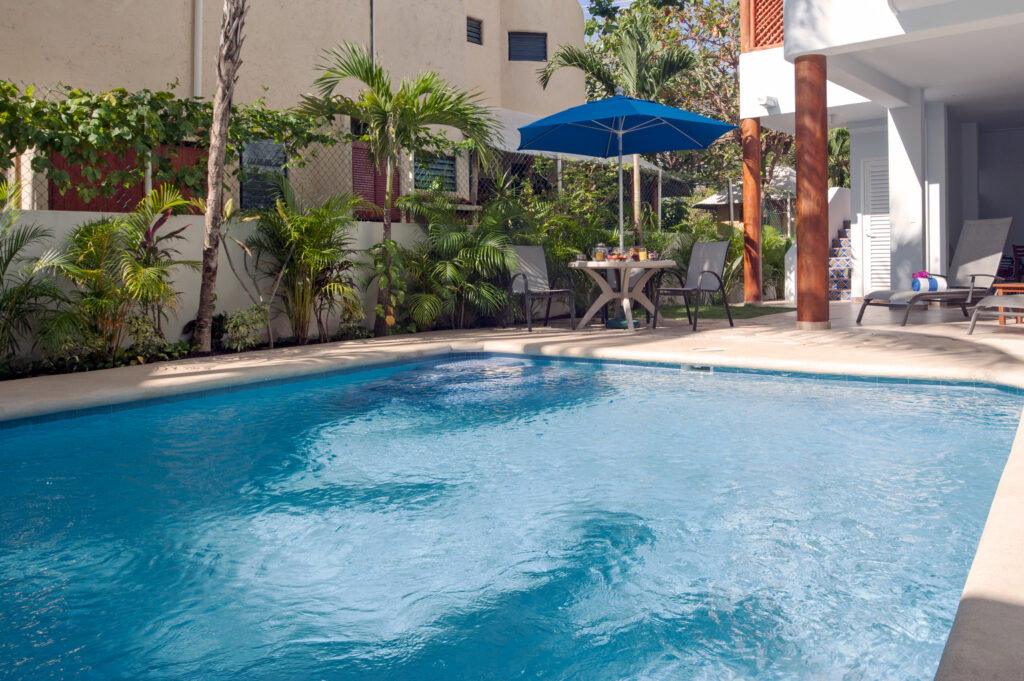 Blue pool bordered by tropical plants and outdoor seating beneath a blue umbrella.