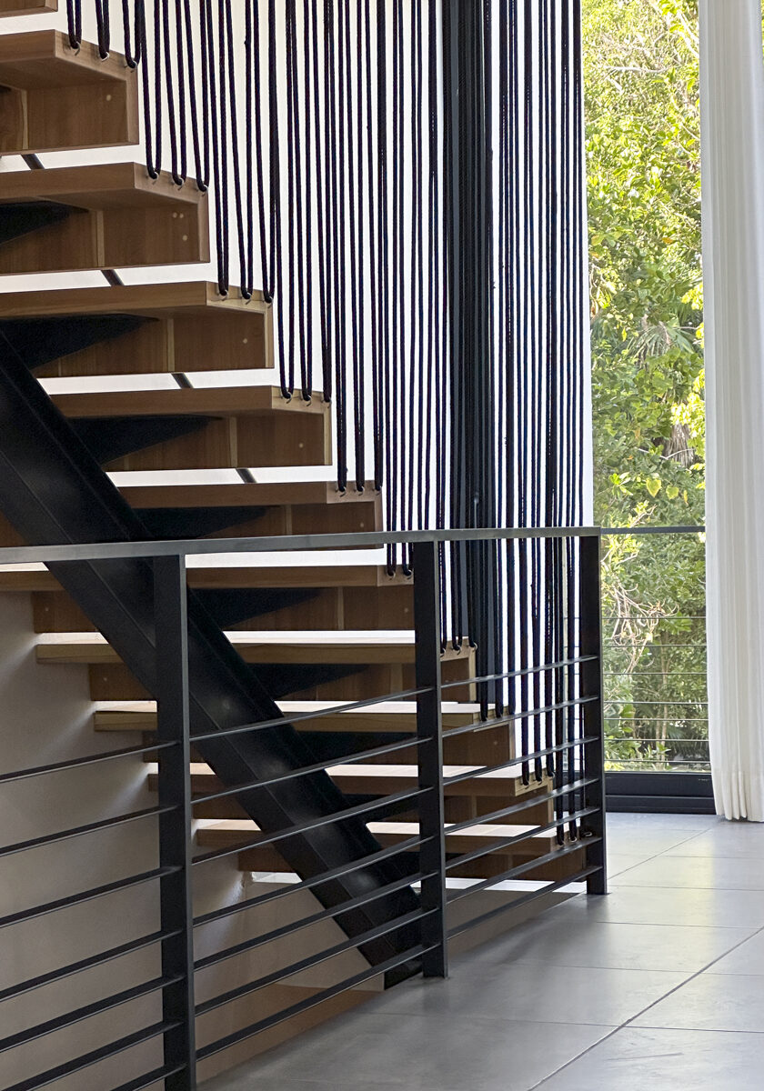 A modern staircase with minimalist wooden steps and sleek black railings, set against a backdrop of large windows that reveal a lush, green forest outside. Light flows into the space, highlighting the clean lines and contemporary design.